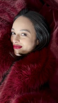 Woman with dark hair and red lipstick wrapped in deep red faux fur, smiling softly while looking sideways.