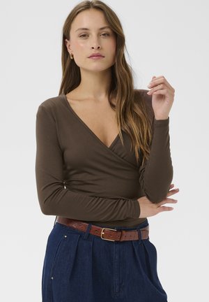 Femme aux longs cheveux bruns portant un top cache-cœur marron à encolure en V, un pantalon bleu taille haute et une ceinture marron, posant les bras croisés.