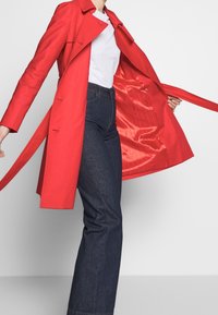 Trench-coat rouge avec une doublure en satin, doté d'une ceinture à la taille et d'un col classique, assorti à un jean denim foncé taille haute.