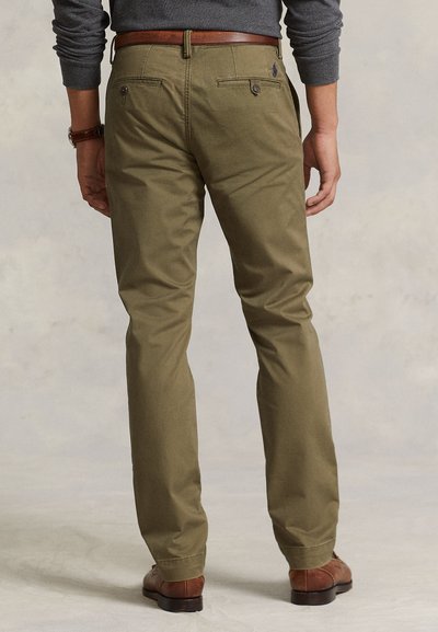 Hombre con pantalones chinos verde oliva, cinturón de cuero marrón, zapatos marrones y camisa de manga larga gris, visto desde atrás y de pie sobre un fondo liso.