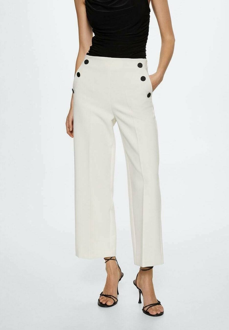 Femme portant un pantalon large taille haute blanc avec des boutons noirs et des sandales à talons noires à lanières, les mains dans les poches.