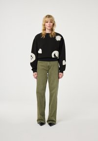 Zwarte sweatshirt met witte grafische accenten, gecombineerd met olijfgroene wijde broek. Gestructureerde stof en een relaxte pasvorm. Zwarte spitse schoenen.