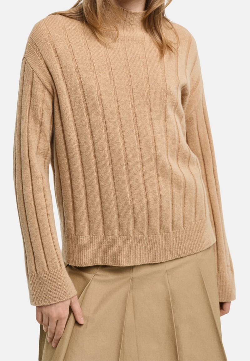 Donna che indossa un maglione a coste beige con maniche lunghe abbinato a una gonna pieghettata color tan su uno sfondo neutro.