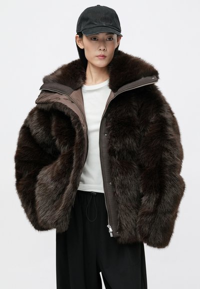 Rudas dirbtinės kailio striukė su gaubtu, su užtrauktuku priekyje, sagų užsegimu ir lygiu vidaus pamušalu. Dėvima ant baltos bluzos ir juodų kelnių.