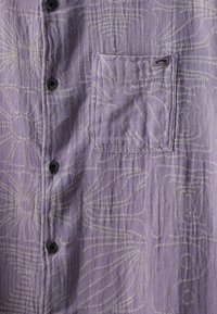 Camisa de botones púrpura con un patrón floral texturizado y un bolsillo en el pecho izquierdo. Los botones negros contrastan con la tela.