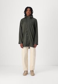 Donkergroen regenjas met een capuchon, voorzien van een drukknoopsluiting aan de voorkant en twee zijzakken, gecombineerd met lichtbeige broek en beige schoenen.
