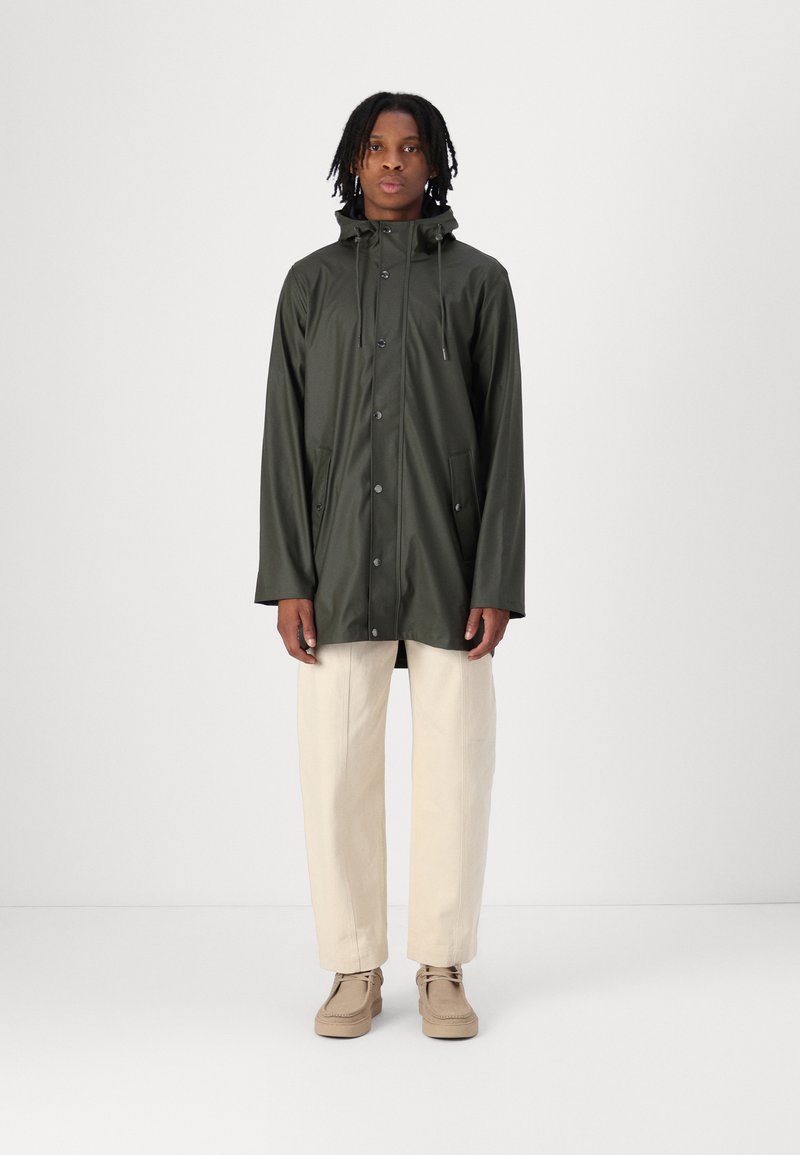 Donkergroen regenjas met een capuchon, voorzien van een drukknoopsluiting aan de voorkant en twee zijzakken, gecombineerd met lichtbeige broek en beige schoenen.