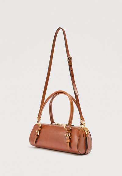 Μικρή καφέ δερμάτινη τσάντα με δύο επάνω λαβές, ρυθμιζόμενο λουρί ώμου, αγκράφες σε χρυσό τόνο και κλείσιμο με φερμουάρ σε απλό φόντο.