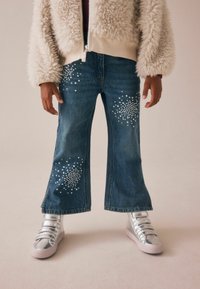 Jeans i denim med utsvängda ben, prydda med silverfärgade rhinestonedekorationer på framsidan. Matchas med glänsande silverfärgade sneakers och en fluffig krämfärgad jacka.