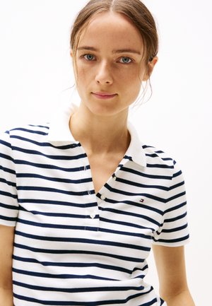 Ung kvinne med lysebrunt hår som har på seg en hvit og marineblå stripete poloshirt med en liten logo på brystet mot en hvit bakgrunn.