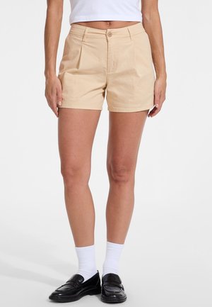 Pantaloni corti in cotone beige con un design sartoriale, tasche frontali e chiusura a un bottone, abbinati a mocassini neri e calzini bianchi.