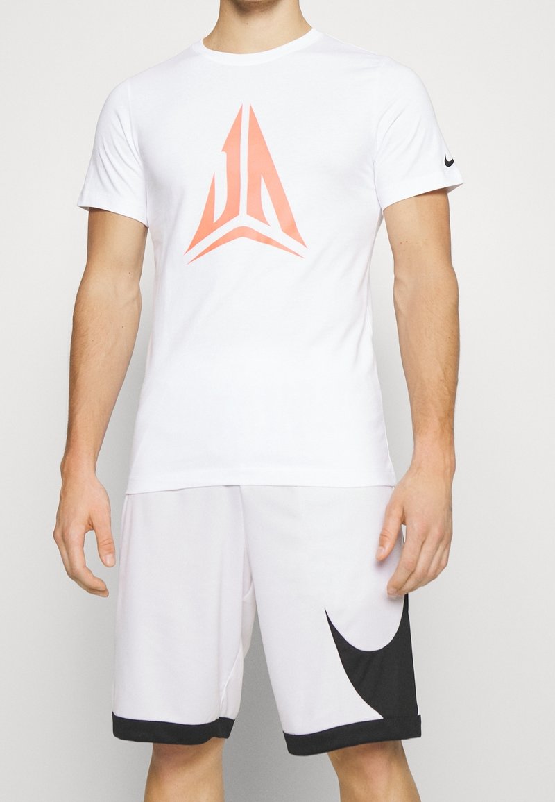 Homme portant une tenue de sport blanche avec un logo géométrique orange sur la chemise et un grand motif swoosh noir sur le short.