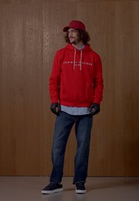 Felpa con cappuccio rossa e logo, camicia blu sotto, jeans blu, guanti neri e sneaker neri, su uno sfondo di legno.