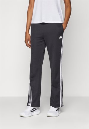 Persoon draagt zwarte trainingsbroek met witte strepen en witte sneakers, staat met één hand in de zak.