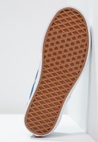 Vita sneakersula med en brun gummisula som har ett strukturerat fiskbensmönster, rundade kanter och blå detaljer på sidan.