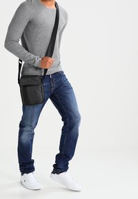 Osoba v šedém tričku s dlouhým rukávem, modrých džínách, bílých teniskách a černé crossbody tašce, která drží popruh levou rukou.
