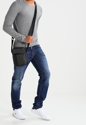 Persona che indossa una maglietta grigia a maniche lunghe, jeans blu, scarpe da ginnastica bianche e una borsa a tracolla nera, tenendo la cinghia con la mano sinistra.