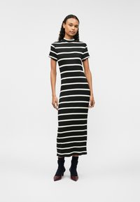 Fekete-fehér csíkos midi ruha bordázott textúrával, rövid ujjakkal és magas nyakkivágással. Sötét zoknival és hegyes orrú magassarkúval viselve.