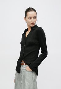 Μαύρο μακρυμάνικο ριμπ πουκάμισο με λαιμόκοψη με σχισμή, κουμπιά μπροστά και στενή εφαρμογή· συνδυασμένο με γκρι μεταλλικό παντελόνι ψηλόμεσο.