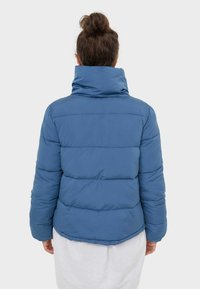 Woman with brown hair in a bun wearing a blue puffer jacket and white skirt, facing away from the camera against a plain background.