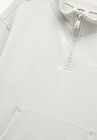 Lys grå, halvmoller pullover lavet af blødt stof, med en frontlomme, minimal broderi og en glat tekstur.