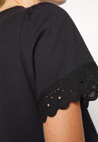 Svart bomullsskjorta med korta ärmar och en scalloped spetskant samt små ögonhål. Slät textur med lätt puff vid axeln.