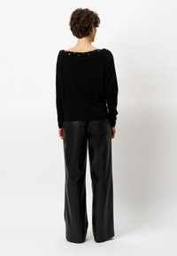 Schwarzer Oversize-Pullover mit weitem Ausschnitt, versehen mit metallischen Akzenten entlang des Rückens. Getragen zu lockeren, hochgeschnittenen schwarzen Lederhosen.