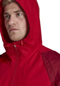 Rode waterdichte jas met capuchon, voorrits en verstelbare toggles. Glad oppervlak met contrasterend donkerrood paneel op de schouder.