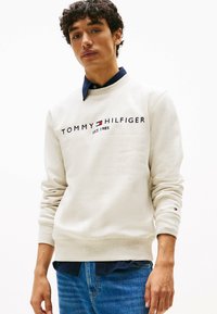 Felpa beige chiaro con polsini e orlo a costine, presenta il logo "TOMMY HILFIGER EST. 1985" in blu scuro e rosso; indossata sopra una camicia blu navy.