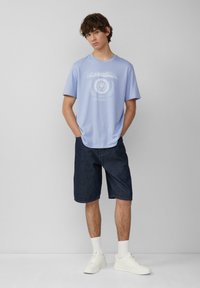 Jasnoniebieska bawełniana koszulka z białym nadrukiem graficznym, w zestawieniu z ciemnoniebieskimi szortami dżinsowymi i białymi sneakersami, na modelce z krótkimi włosami.