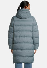 Frau mit dunklem Haar in einem Pferdeschwanz, die eine lange, graue, gesteppte Daunenjacke mit einer großen Kapuze trägt, mit dem Rücken zu einem schlichten Hintergrund gewandt.