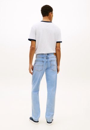 Mężczyzna stojący tyłem, ubrany w białą koszulkę z ciemnym obszyciem, jasnoniebieskie dżinsy i czarno-białe trampki na jednolitym tle.