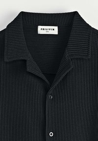 Chemise noire à côtes avec col pointu, dotée de boutons sur le devant. Le tissu semble doux et texturé avec des rayures verticales. Étiquette de taille visible.