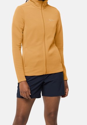 Kvinde iført en sennepsgul ribbet lynlåstrøje og marineblå shorts, stående med armene afslappet ned langs siderne.