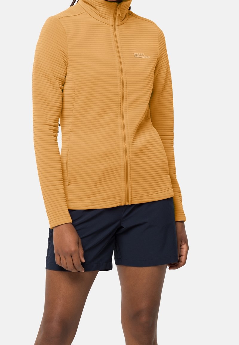 Femme portant une veste zippée côtelée jaune moutarde et un short bleu marine, debout, les bras détendus le long du corps.