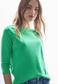 Grünes Langarmshirt aus einem weichen Stoff mit einer lockeren Passform. Rundhalsausschnitt und leicht taillierter Saum. Kombiniert mit blauen Jeans.