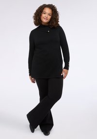 Svart stickad tröja med hög krage, med en knappdetalj vid halsen och gulddetaljer på ärmarna, ihop med svarta utsvängda byxor.