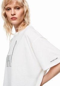T-shirt bianca oversize con maniche corte, caratterizzata da una stampa grafica sul davanti e testo sulla manica. Realizzata in morbido cotone.