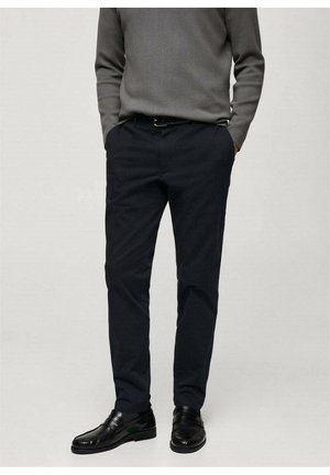 Homme portant un pantalon noir sur mesure, des chaussures habillées en cuir noir et un pull gris à manches longues, les mains dans les poches.