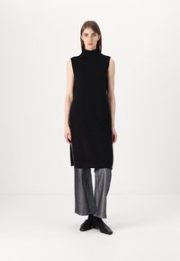 Μαύρο μίνι φόρεμα χωρίς μανίκια με ψηλό λαιμό, που διαθέτει πλάγιες σχισμές. Συνδυασμένο με ανοιχτό γκρι παντελόνι σε φαρδιά γραμμή και μαύρα παπούτσια.