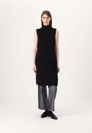 Schwarzes ärmelloses Strickkleid mit hohem Kragen, das seitliche Schlitze aufweist. Kombiniert mit hellgrauen weit geschnittenen Hosen und schwarzen Schuhen.