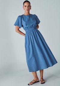 Modra denim obleka s kratkimi napihnjenimi rokavi, elastičnim pasom in krilom do gležnjev. Gladka textura in minimalističen dizajn brez vidnih vzorcev.