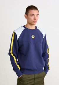 Námornícky modrý crewneck mikina s svetlomodrými ramennými panelmi a žltými bočnými akcentmi. Na hrudi sa nachádza hexagonálne logo Nike.