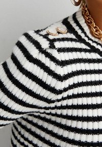 Pull à rayures noires et blanches en fil doux ; présente une texture côtelée et des boutons décoratifs sur l'épaule.