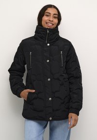 Žena usmievajúca sa, oblečená v čiernej prešívanej bunde s vysokým golierom, zipsom a patentkami, v kombinácii so svetlomodrými džínsami, stojí proti bielej stene.