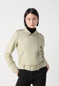 Jachetă din faux piele verde deschis, cu fermoar frontal, detalii cu curea și accesorii din argint. Dispune de un guler structurat și buzunare laterale.