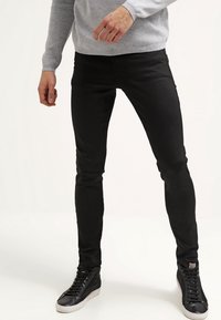 Jean slim noir en denim, avec une texture lisse et un design minimaliste. Associé à des baskets noires et un pull gris.