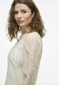 Lekka, kremowa bluzka z teksturowanymi rękawami i zawiązanymi detalami. Miękka tkanina o płynnej, eleganckiej sylwetce.