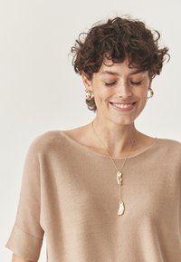 Beżowy sweter z krótkim rękawem o gładkiej fakturze. Złota naszyjnik z dwoma organicznymi zawieszkami zwisa poniżej dekoltu.