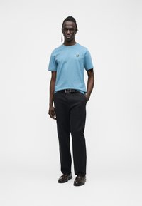 Light blue cotton t-shirt with a round neck and yellow emblem, paired with black trousers and dark brown shoes, worn by a standing male model.